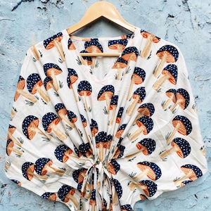 May include: A white, V-neck kaftan with a repeating pattern of blue-capped mushrooms with orange gills and tan stems. The kaftan is hanging on a wooden hanger against a blue wall. The kaftan has a tie closure at the waist.