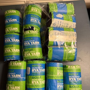 May include: A collection of Bucilla Deluxe Quality Rya Yarn skeins wrapped in green and blue paper labels. Each label features the text Bucilla Deluxe Quality Rya Yarn, 60% pure wool, 40% nylon, and permanently mothproof. The yarn is packaged in plastic.