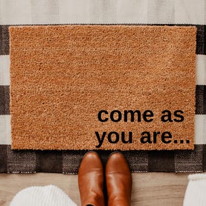 May include: A rectangular doormat with the text "come as you are..." in black lettering. The doormat is placed on a black and white checkered rug. Brown boots and blue jeans are visible.