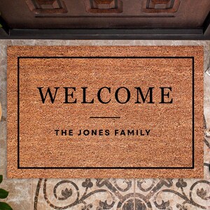 May include: A rectangular doormat made of natural coir fiber. The mat features the word "WELCOME" in a bold, black font, with "THE JONES FAMILY" below. A black border frames the text.