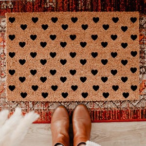 May include: A rectangular doormat made of natural coir fiber, featuring a repeating pattern of black hearts. The mat is placed on a patterned rug with shades of red, brown, and cream. Brown boots are visible at the bottom.