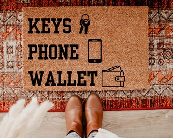 May include: A rectangular doormat made of natural coir fiber, featuring the words "KEYS PHONE WALLET" in bold black letters. The design includes key, phone, and wallet icons. The mat is placed on a patterned rug, with a person's feet and boots visible.