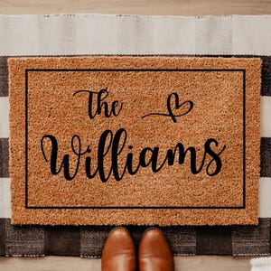 May include: A rectangular coir doormat with the family name "The Williams" in black script, with a heart symbol. The mat is framed in black and sits on a black and white checkered rug. Brown shoes are visible at the bottom.