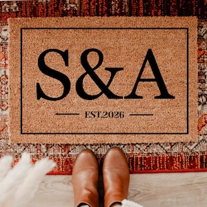 May include: A rectangular doormat with the letters "S&A" in bold black font, with "EST.2026" below. The mat is a light brown color and is framed by a black border. The mat is placed on a patterned rug.
