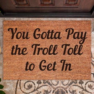 May include: A rectangular doormat made of a natural fiber material, featuring the phrase "You Gotta Pay the Troll Toll to Get In" in a bold, black script. The mat is placed in front of a door.