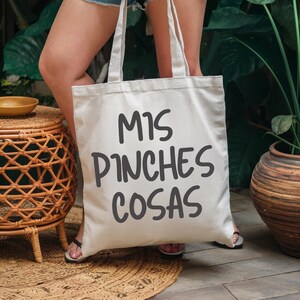 May include: Cream-colored tote bag with the phrase "MIS PINCHES COSAS" in dark gray lettering. The bag has two long handles and is held in front of a person. The background includes greenery and a woven side table.