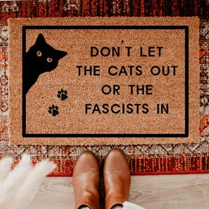May include: A rectangular doormat with a black border and a light brown surface. The mat features a black cat silhouette in the upper left corner, paw prints, and the text "DON'T LET THE CATS OUT OR THE FASCISTS IN" in black.