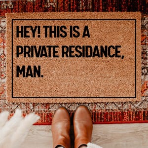 May include: A rectangular doormat with the text "HEY! THIS IS A PRIVATE RESIDANCE, MAN." in black letters. The mat is brown and framed in black. The mat is on a patterned rug with brown leather boots visible.