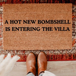 May include: A rectangular coir doormat with the text "A HOT NEW BOMBSHELL IS ENTERING THE VILLA" in black. The mat is placed on a patterned rug, with brown boots and denim jeans visible.