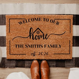 May include: A rectangular doormat with the text "WELCOME TO OUR HOME THE SMITHS FAMILY EST 2026" in black script. The mat is brown with a black border and sits on a black and white checkered rug. Brown boots are visible.