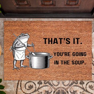 May include: A rectangular doormat with a frog chef illustration and the text "THAT'S IT. YOU'RE GOING IN THE SOUP." The mat is brown with a textured surface. A frog in a chef's apron stirs a pot.