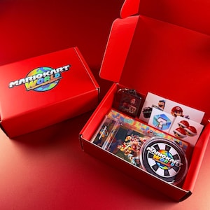 Op de afbeelding: Rode kartonnen doos met het "MARIOKART WORLD" logo op het deksel. Binnenin zijn diverse thematische items zichtbaar, waaronder stickers, een sleutelhanger en een onderzetter. De doos is open en onthult de inhoud.