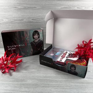 May include: A Silent Hill gift box, open to show contents. The box features a portrait of a young woman in a school uniform. Inside are trading cards, a comb, and other items. The box is black with red and white accents.