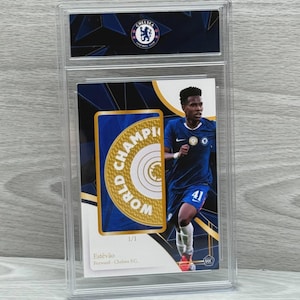 Puede incluir: Una tarjeta coleccionable de Estêvão del Chelsea FC, dentro de un protector. La tarjeta muestra al jugador con uniforme azul, con las palabras "WORLD CHAMPION" y el logo del Chelsea. La tarjeta está marcada 1/1.