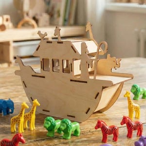 Puede incluir: Un arca de juguete de madera con una variedad de figuras de animales coloridas, incluyendo elefantes, jirafas, leones, cebras, monos e hipopótamos. El arca está hecha de madera clara y tiene un diseño detallado. Los animales son de varios colores.