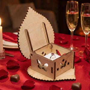 Puede incluir: Un joyero de madera con un corte de mariposa y un anillo dentro, sobre un mantel rojo con pétalos de rosa, bombones y copas de champán. Velas encendidas y rosas crean un ambiente romántico.