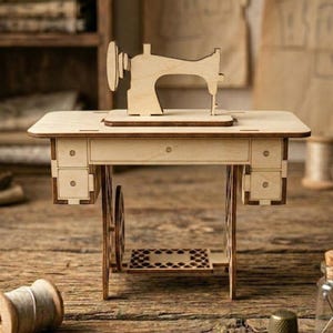 K&ouml;nnte beinhalten: Ein Miniatur-N&auml;hmaschinenmodell aus hellem Holz mit Tisch, Schubladen und Fu&szlig;pedal. Das Modell steht auf einer Holzoberfl&auml;che. Eine Garnrolle, ein Fingerhut und eine kleine Glasflasche mit Nadeln sind ebenfalls vorhanden.