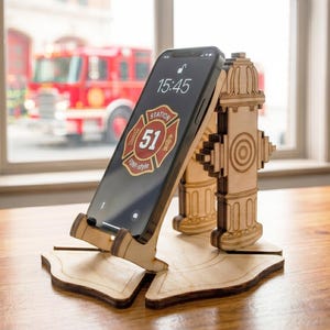 May include: A wooden phone stand shaped like a fire hydrant, holding a smartphone. The phone displays the time and a Station 51 logo. The stand is made of light-colored wood and sits on a wooden surface, with a fire truck visible in the background.