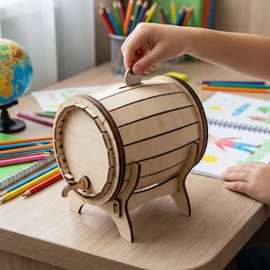 Pode incluir: Um cofre de madeira clara em forma de barril, com uma ranhura para moedas e uma torneira. O cofre est&aacute; numa secret&aacute;ria com um globo, l&aacute;pis de cor e a m&atilde;o de uma crian&ccedil;a a inserir uma moeda.