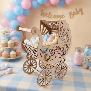 May include: A wooden baby carriage filled with pastel-colored treats, including marshmallows and baby shoes. The carriage has intricate cut-out designs and sits on a blue and white checkered tablecloth. Balloons and a banner that says "Welcome Baby" are in the background.