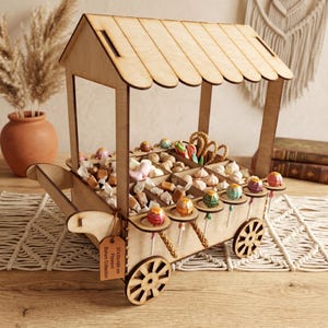 Peut inclure: Un chariot à bonbons en bois rempli de divers bonbons, dont des sucettes, des caramels et des guimauves. Le chariot a un auvent et des roues à rayons. Une étiquette indique "37x30x40 cm Plywood, Bohem Collection."