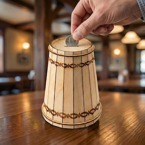 Può includere: Una salvadanaio a forma di botte in legno chiaro con motivi decorativi marrone scuro. Una mano sta inserendo una moneta nella fessura. Il salvadanaio è appoggiato su un tavolo di legno.