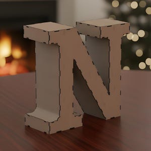 May include: A large, free-standing letter "N" made of light brown cardboard. The letter has a boxy, geometric design with visible cut lines. It is placed on a dark wooden surface, with a blurred fireplace and Christmas tree in the background.
