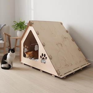 Puede incluir: Una casa para gatos de madera clara con forma de casa, con recortes en forma de huellas y siluetas de gatos grabadas. Un gato naranja descansa dentro, mientras que un gato blanco y negro est&aacute; sentado cerca.