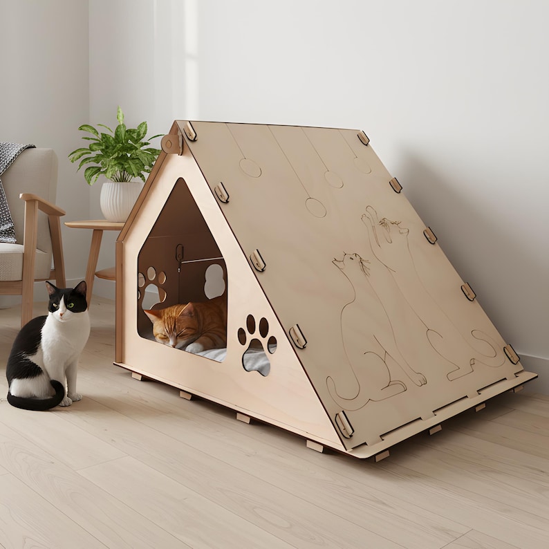 Puede incluir: Una casa para gatos de madera clara con forma de casa, con recortes en forma de huellas y siluetas de gatos grabadas. Un gato naranja descansa dentro, mientras que un gato blanco y negro est&aacute; sentado cerca.
