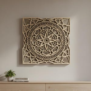 Puede incluir: Una pieza de arte mural cuadrada de madera en capas con un diseño de mandala detallado. La obra de arte presenta un patrón floral simétrico con intrincados recortes, mostrando un color de madera natural. La pieza está montada en una pared de tonos neutros.
