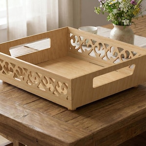 Puede incluir: Una bandeja de madera clara con patrones decorativos recortados en los lados. La bandeja tiene asas y está sobre una mesa de madera. Un jarrón con flores está en el fondo.