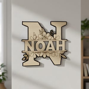 Puede incluir: Arte de pared de madera con el nombre "NOAH" dentro de una letra "N" decorada con flores. El recorte de madera tiene un color de madera natural con contorno negro y está montado en una pared blanca. El diseño floral incluye patrones detallados de flores y hojas.