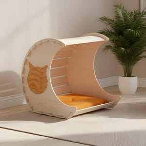 Puede incluir: Una cama para gatos de madera clara en forma de media luna. La cama tiene un coj&iacute;n naranja y un recorte de cara de gato en el lateral. La cama est&aacute; hecha de madera clara y tiene un dise&ntilde;o moderno.