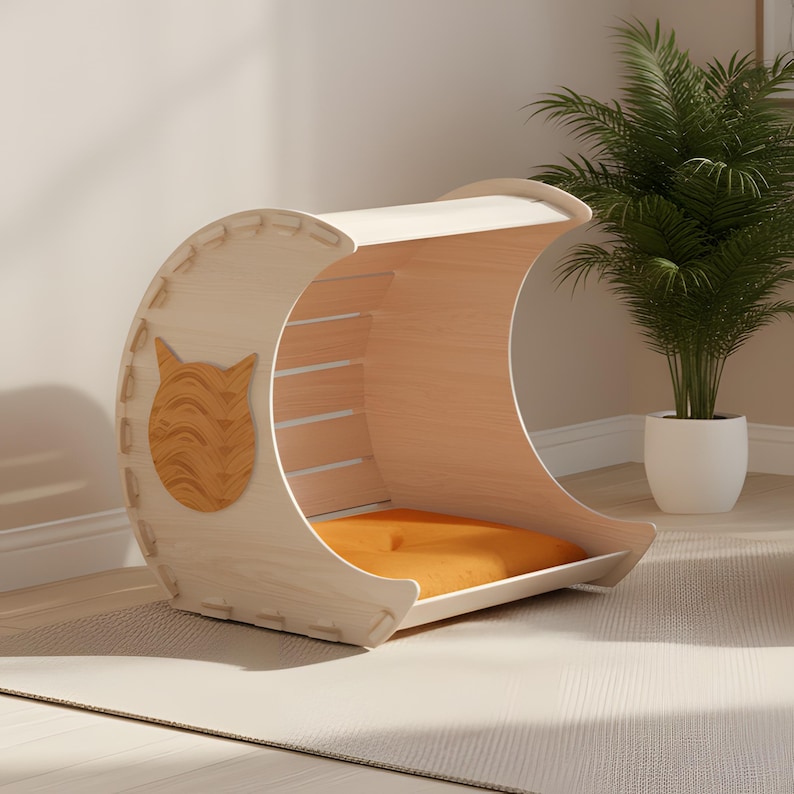 Puede incluir: Una cama para gatos de madera clara en forma de media luna. La cama tiene un coj&iacute;n naranja y un recorte de cara de gato en el lateral. La cama est&aacute; hecha de madera clara y tiene un dise&ntilde;o moderno.