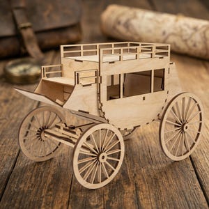 May include: A wooden stagecoach model with intricate details, including spoked wheels, a passenger compartment, and a driver's seat. The model is made of light-colored wood and sits on a wooden surface.