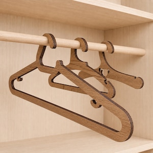 Può includere: Tre grucce di legno con un motivo a venature naturali sono appese a un'asta di legno all'interno di un armadio di legno colorato chiaro. Le grucce hanno un design semplice e moderno con spalle curve e un gancio per appendere i vestiti.