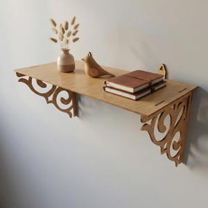 Puede incluir: Una estantería de pared de madera clara con soportes decorativos de volutas. La estantería contiene un jarrón con flores secas, una figura de pájaro de madera y dos cuadernos encuadernados en cuero marrón. La estantería está montada en una pared gris claro.