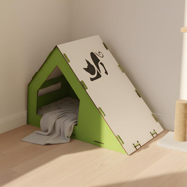 Puede incluir: Una moderna caseta para gatos de forma triangular, con marco verde y techo blanco. El techo presenta una silueta de gato negro. En el interior, una manta gris descansa sobre una cama suave. La caseta es de madera.