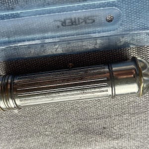May include: A cylindrical silver-toned metal torch with a ribbed texture along the body and a rounded glass lens at one end, positioned next to a clear plastic ruler that shows the object is approximately 11.5 cm in total length.
