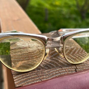 Op de afbeelding: Vintage cat-eye bril met een zilveren montuur en heldere glazen. De monturen hebben decoratieve geëtste details op de slapen. De glazen zijn lichtgeel van kleur. De bril rust op een houten oppervlak.