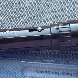 Könnte beinhalten: Eine dunkelbraune zylindrische Taschenlampe mit geriffelter Endkappe und einem seitlichen Metallschiebeschalter. Die Taschenlampe liegt über einem transparenten Kunststofflineal, das eine Länge von etwa 11,5 Zentimetern anzeigt.