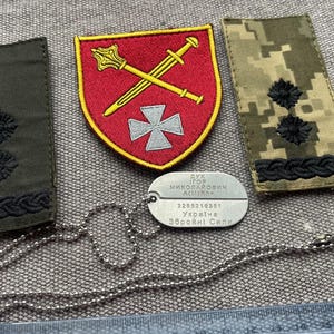 Puede incluir: Dos hombreras militares rectangulares, una de color verde oliva sólido y otra con camuflaje digital, cada una con dos estrellas negras bordadas y un borde trenzado. Un parche rojo en forma de escudo con una maza y una espada doradas.