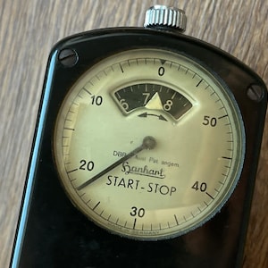 May include: Close-up of a vintage black stopwatch with a cream-colored face. The face features black numerals, a needle, and the text "Sanhart START-STOP". A small dial indicates the time. The stopwatch is set against a wooden surface.