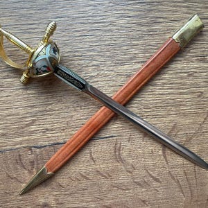 Könnte beinhalten: Ein Brieföffner in Form eines Miniaturschwerts mit goldenem Griff, einem weiß-goldenen Zierknauf und einer silberfarbenen Klinge. Er wird von einer hellbraunen Scheide mit goldfarbenen Metallakzenten an Spitze und Öffnung begleitet.