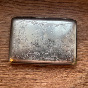 Peut inclure: Un étui à cigarettes rectangulaire en métal argenté avec une gravure détaillée représentant un soldat tenant un fusil, la tour du Kremlin et un bouclier portant l'inscription 30 LET ZA OCHRANE CCCP. La surface présente une finition polie.