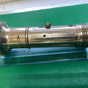 May include: A vintage silver-toned flashlight with a ribbed end and a round, textured head. The flashlight has a small switch and a brass-colored button on top. It is lying on a green surface next to a ruler.