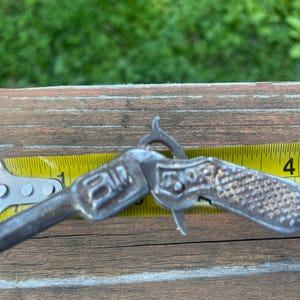May include: A small silver-toned metal toy pistol with a textured grip and a hammer mechanism, positioned against a yellow measuring tape that shows the object is approximately 3.5 inches in length from the barrel tip to the handle end.