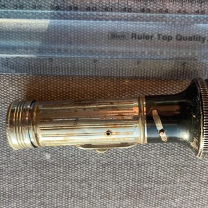 Könnte beinhalten: Eine Vintage-Taschenlampe aus Metall mit geriffeltem, silberfarbenem Gehäuse und schwarzem Kopf. Die Taschenlampe liegt unter einem transparenten Kunststofflineal und misst etwa 11,5 cm von der Basis bis zum vorderen Linsenrand.