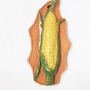 May include: A ceramic corn on the cob wall hanging with green leaves and a yellow cob. The corn is on a peach-colored background with a textured surface.