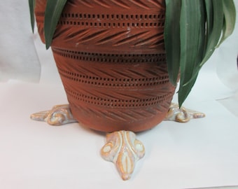 May include: A terracotta planter with a decorative design and a white ceramic base with three feet. The planter is decorated with a pattern of small holes and lines.